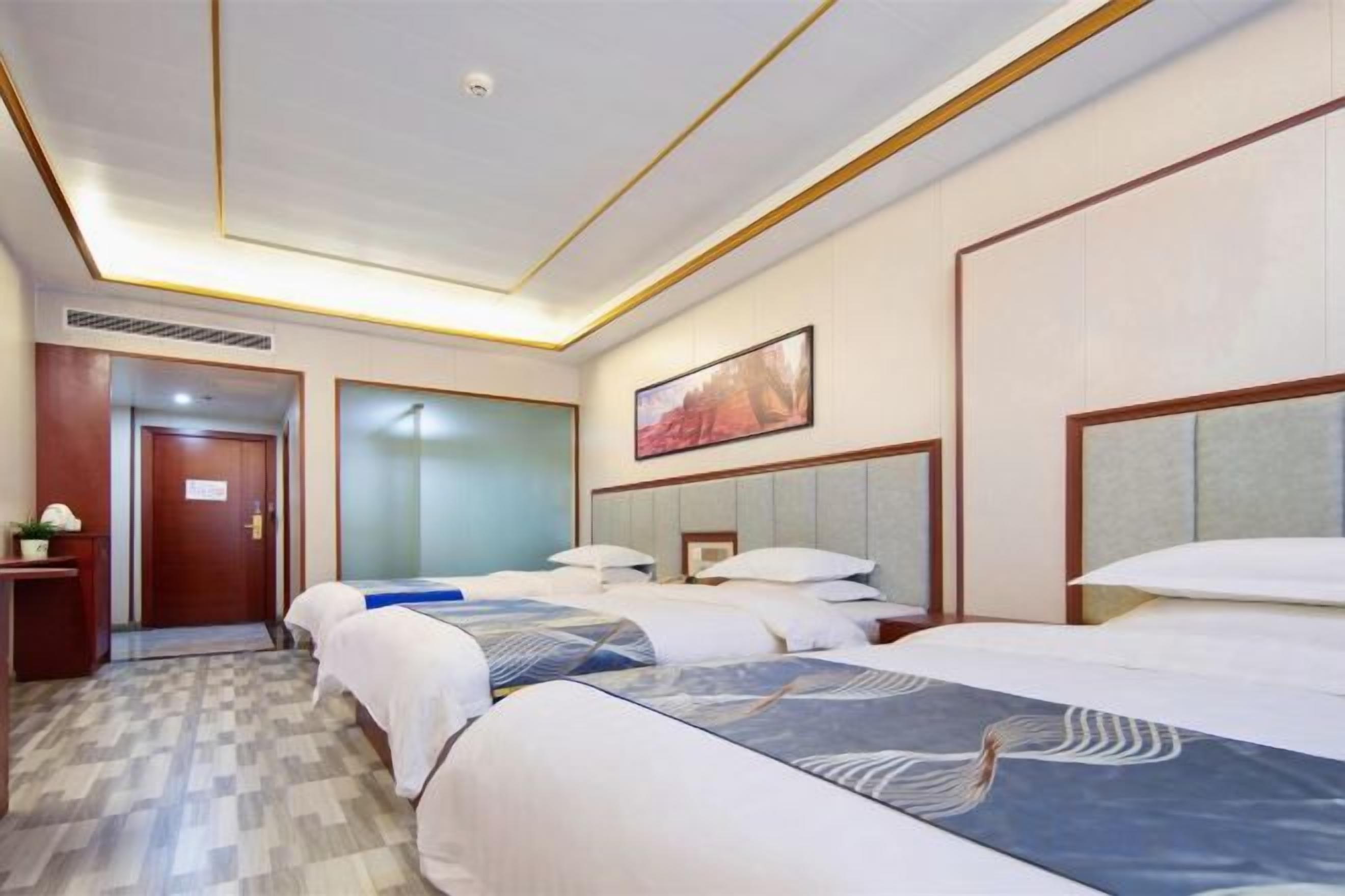 A hotel room with three beds, a door, a painting, and a small table.