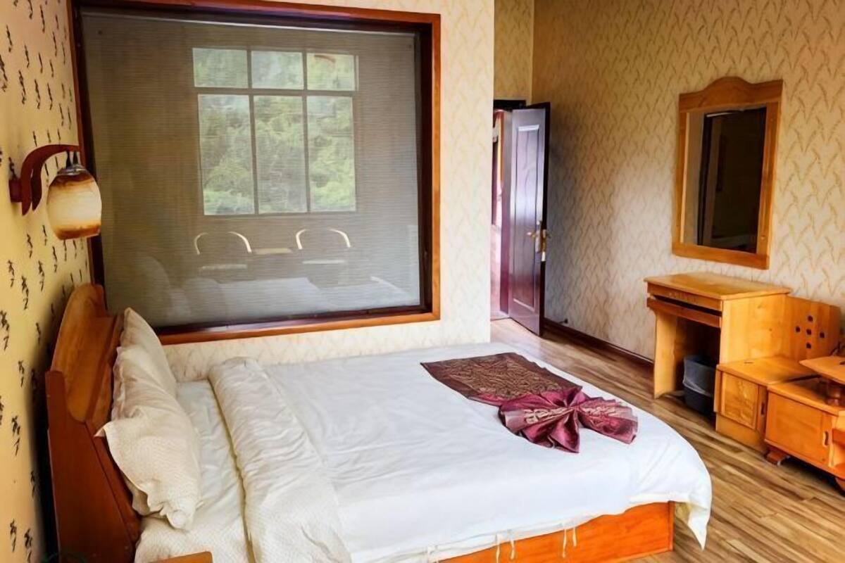 A bedroom with a large bed, a desk, a chair, a wardrobe, and a window with a view of trees.