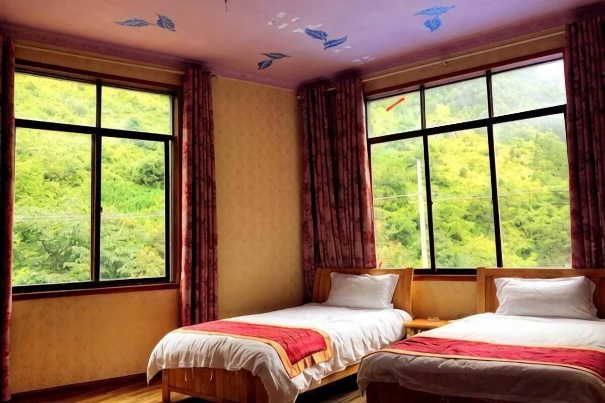 A room with two beds, large windows with curtains, and a view of greenery outside.