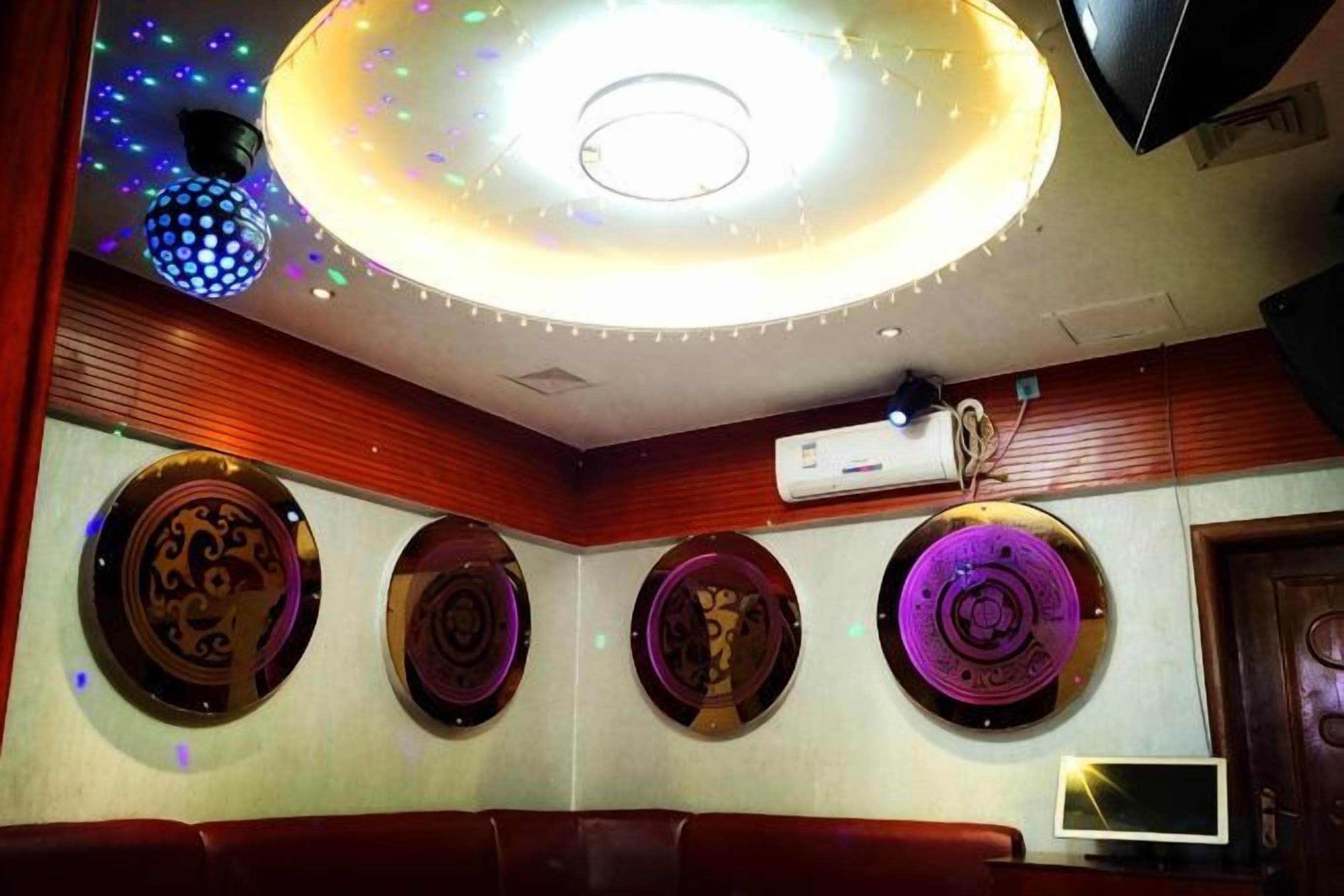 A room with a circular ceiling light, wall-mounted decorative plates, a flat-screen TV, and a wall-mounted air conditioning unit.