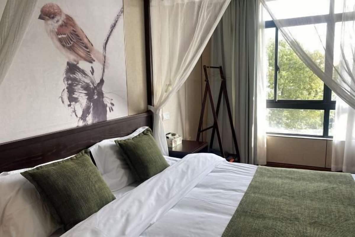 A neatly made bed with white and green bedding, a painting of a bird on the wall, and a window with curtains.