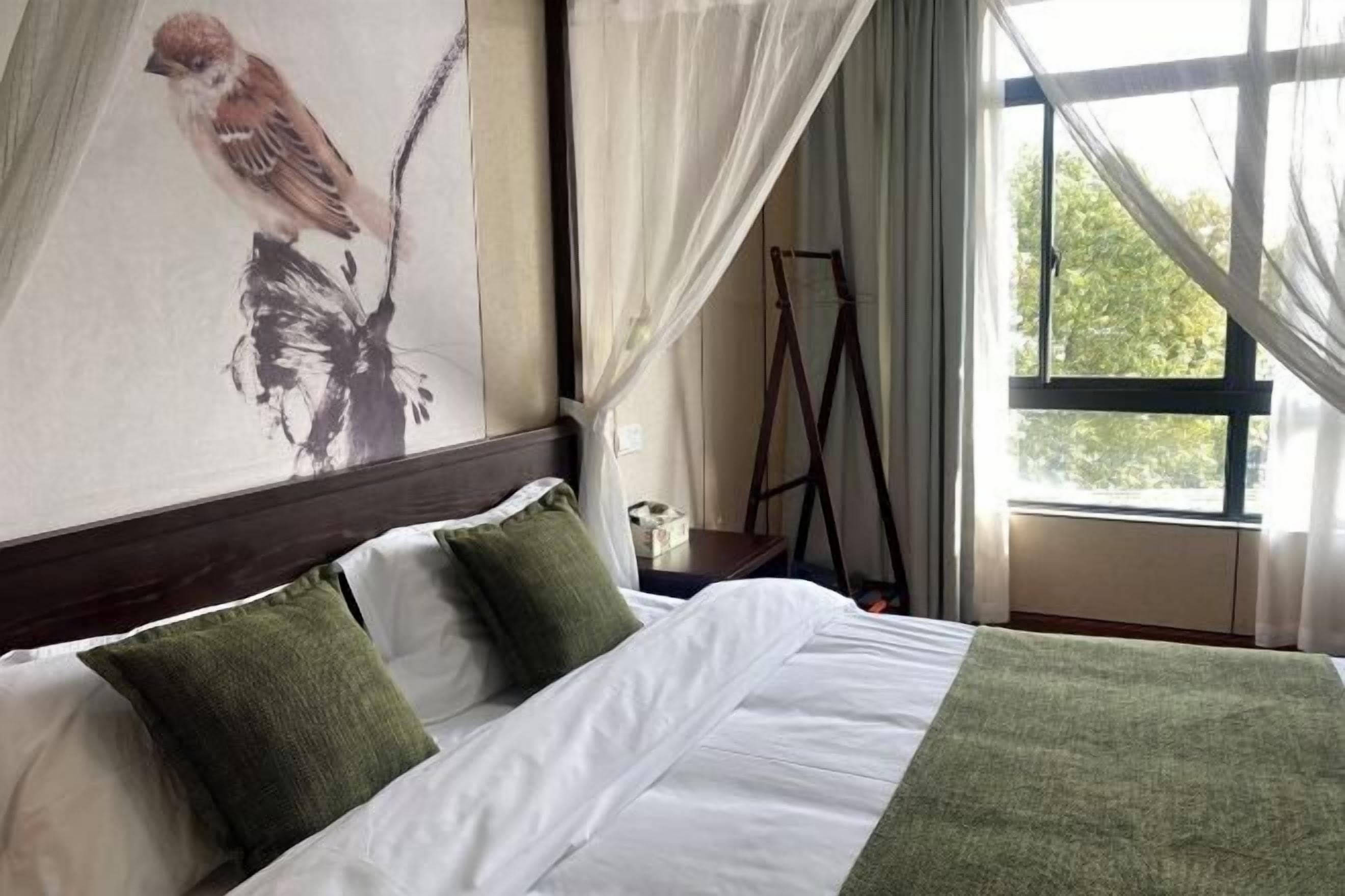 A hotel room with a bed, green pillows, a painting of a bird, and a window with curtains.