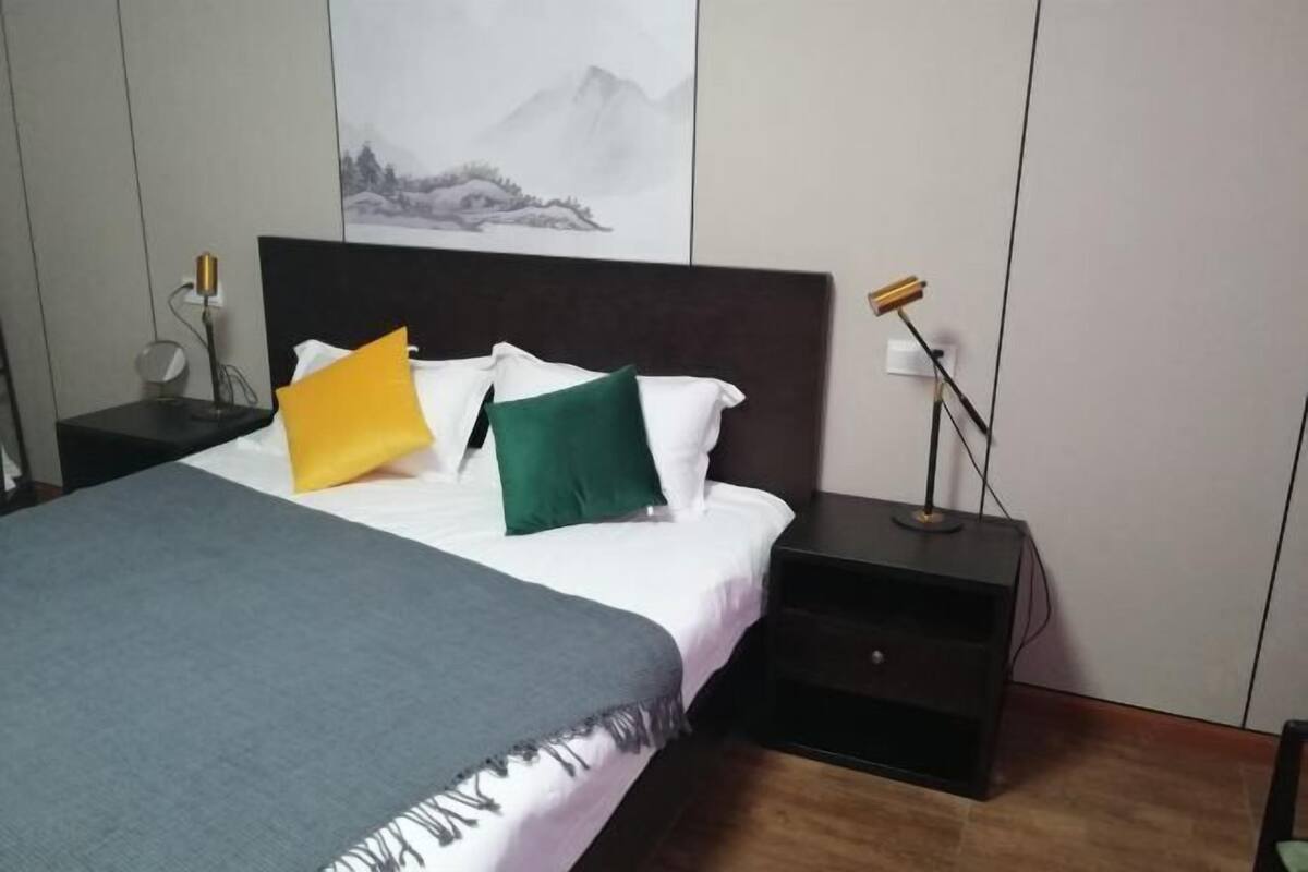 A neatly made bed with a grey blanket, white pillows, and two decorative pillows in yellow and green. There is a nightstand with a lamp and a power outlet on the wall.
