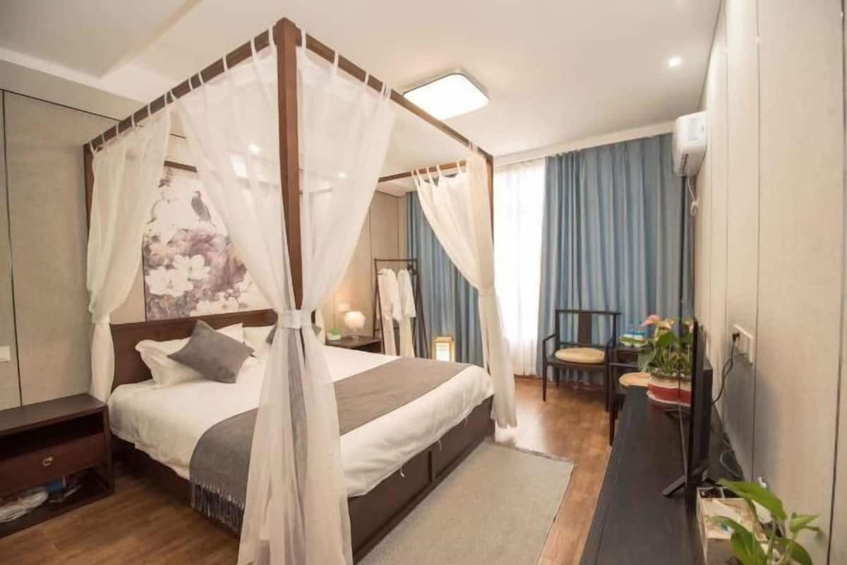 A bedroom with a four-poster bed, a bedside table, a chair, a TV, and a potted plant.