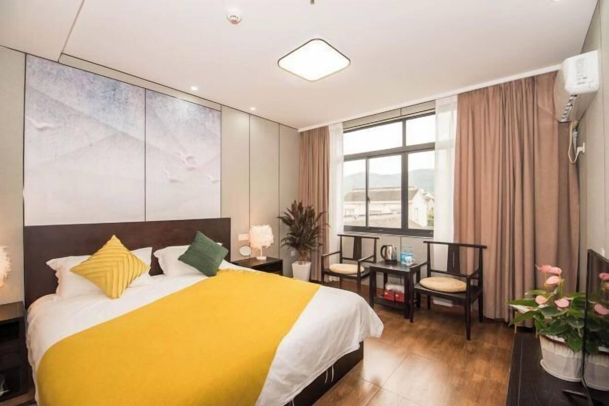 A hotel room with a bed, a desk, a chair, a potted plant, and a window with curtains.