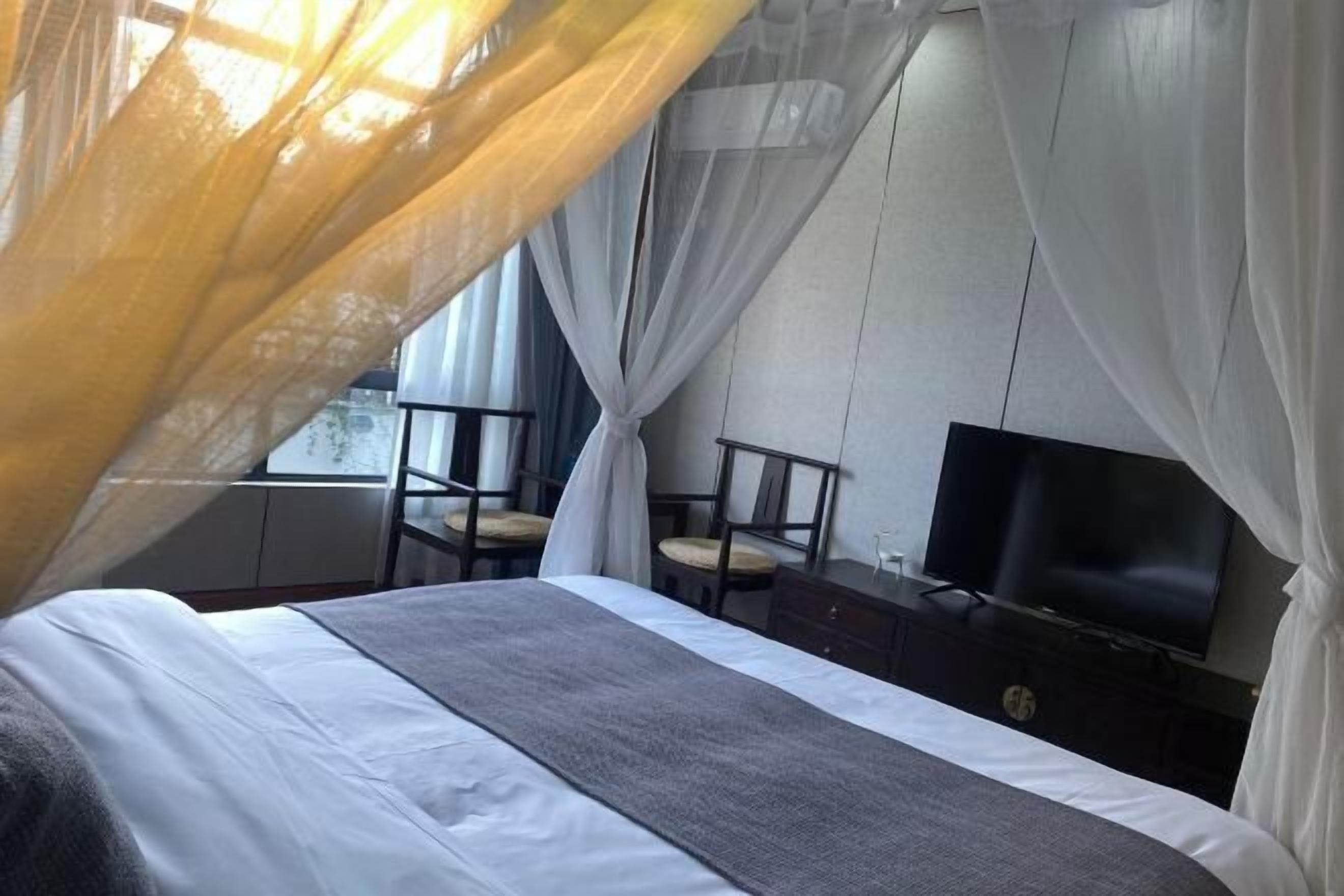 A bedroom with a bed, a TV, and a window with sheer curtains.