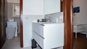 Een moderne badkamer met een wit wastafelmeubel, spiegel en aanrecht. Er staat een prullenbak en een wasmand in de hoek.