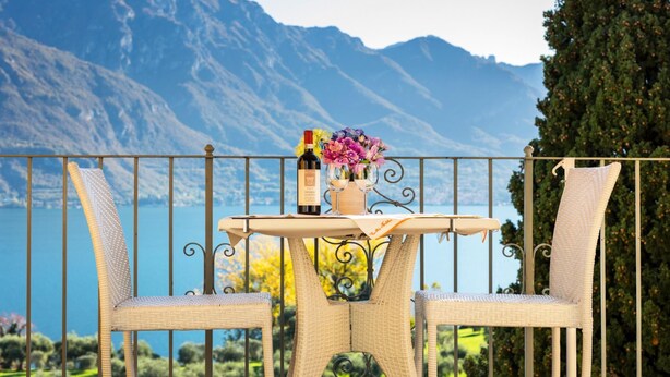 Een balkon met rieten stoelen, een tafel met een fles en glazen, en uitzicht op bergen en een meer.