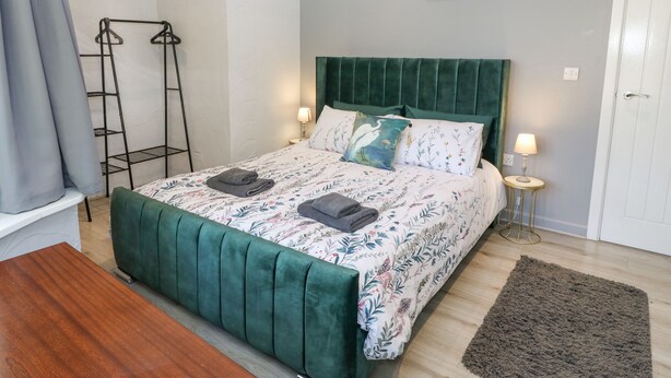 Una cama bien hecha con cabecera verde, cubrecama estampado con flores y dos almohadas. Hay dos lámparas de noche sobre mesitas, una alfombra gris en el suelo y un perchero de metal contra la pared.