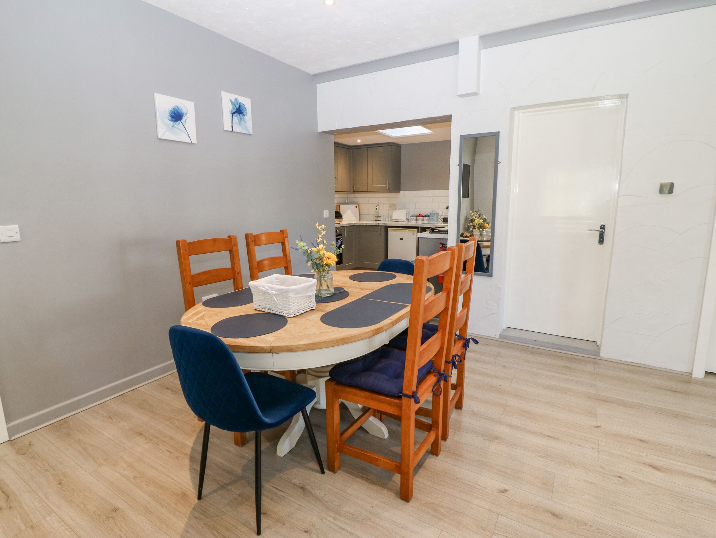 Un comedor con una mesa redonda, seis sillas de madera y una silla con cojín azul. Se observan dos cuadros de flores colgados en la pared y una puerta blanca al fondo.