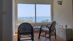 Een kamer met een groot raam dat uitzicht biedt op de zee en palmbomen. Er staan twee rieten stoelen en een klein tafeltje met een glas op.