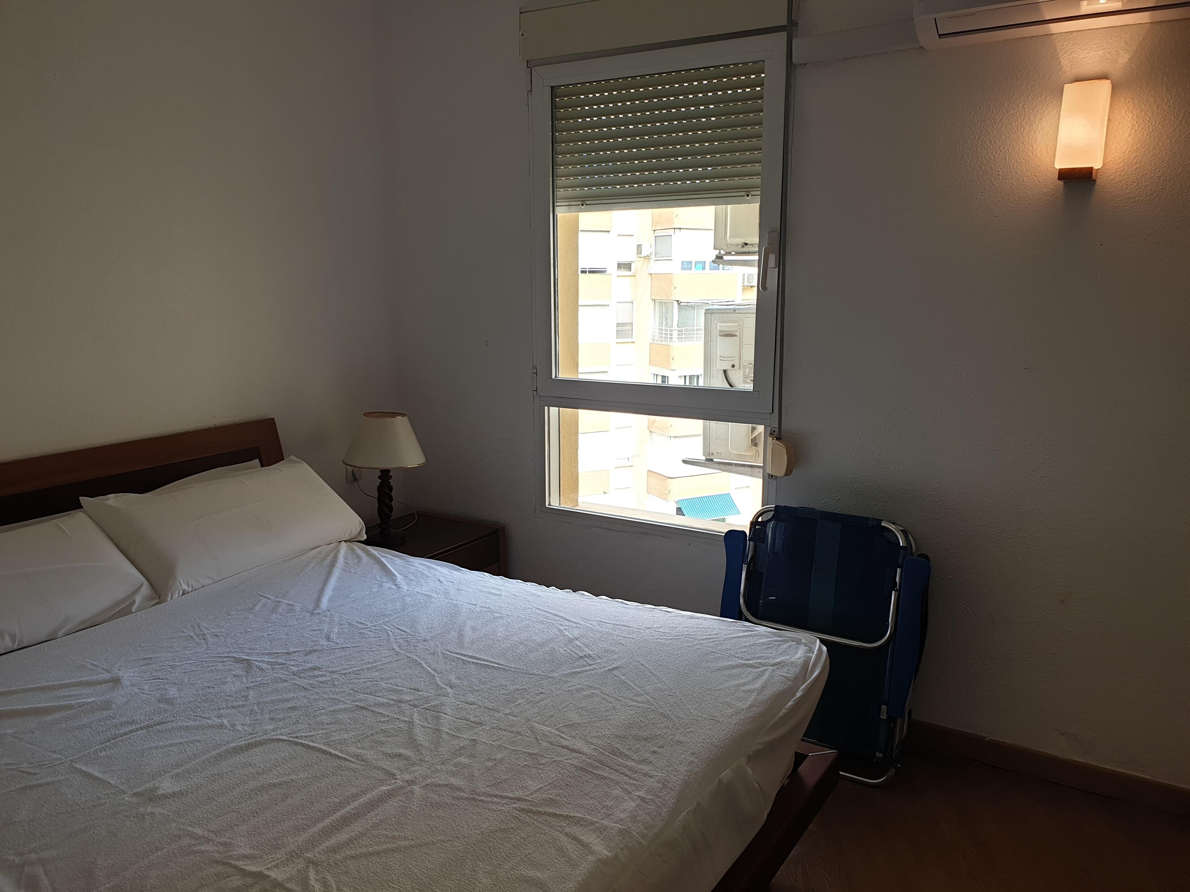 Una habitación de hotel con cama, mesita de noche con lámpara, ventana con persianas y maleta.