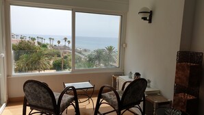 Een kamer met twee stoelen en een kleine tafel, een groot raam met uitzicht op het strand en de oceaan, en een wandlamp.