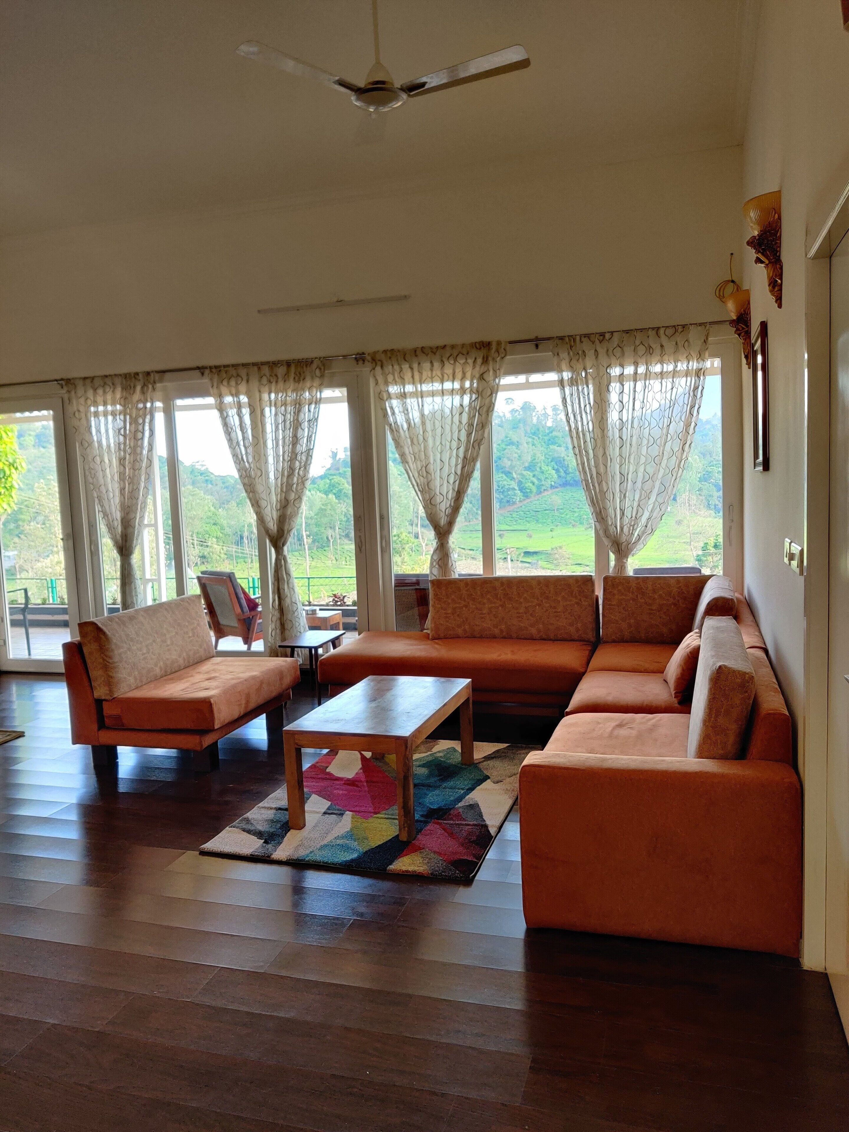 A spacious living room with a large sofa set, a coffee table, and a ceiling fan. Large windows with sheer curtains allow natural light and views of greenery outside.