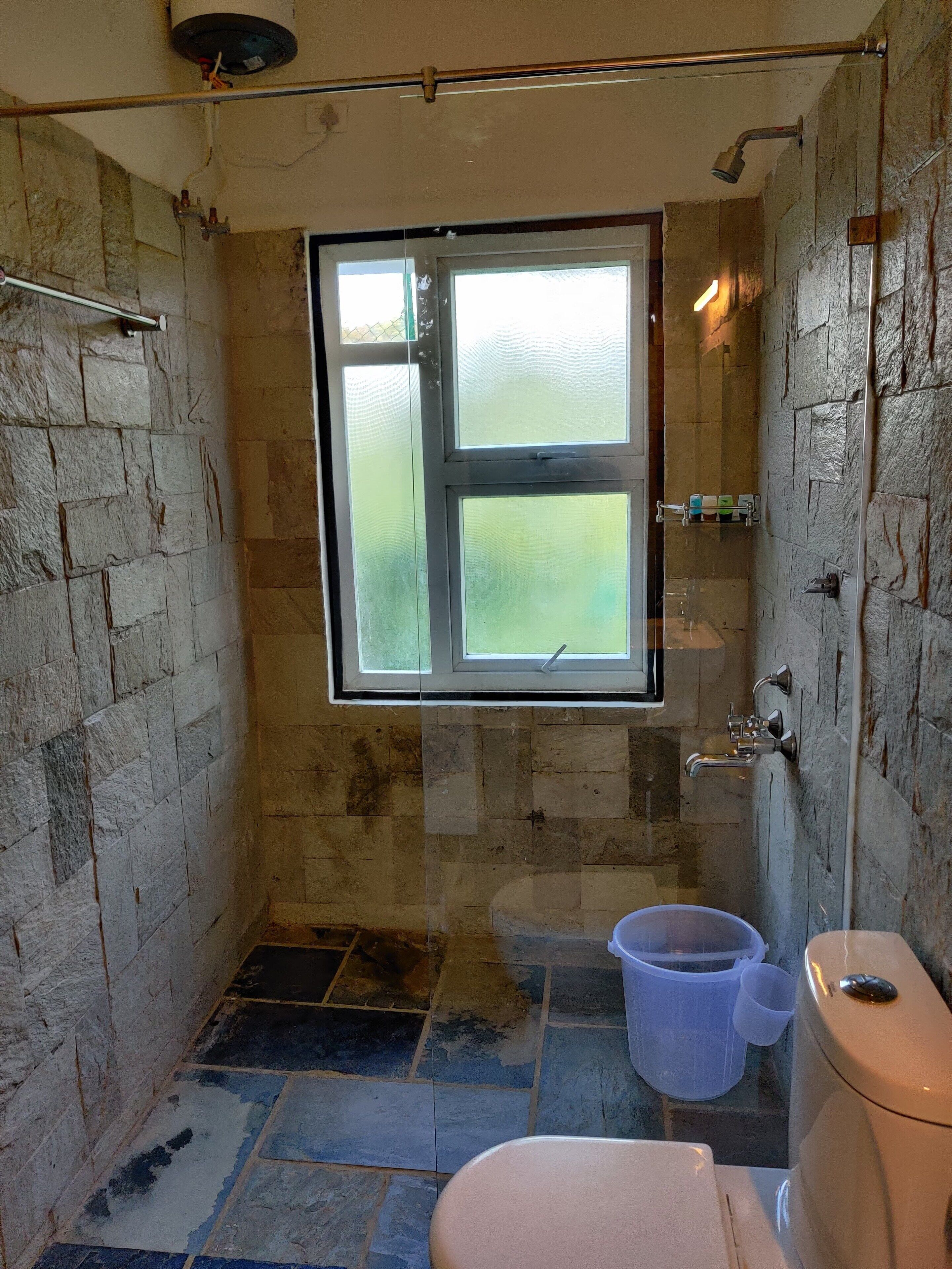 Une petite salle de bain aux murs de pierre, avec une fenêtre, des toilettes, une douche et un seau.
