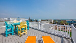 A wooden deck with outdoor furniture, a white railing, and a view of the ocean and beach houses. - Located in the sought-after gated Village Walk community is a charming tropical (Port Aransas)