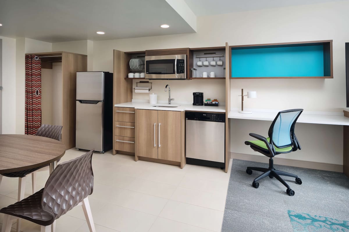 A compact kitchen with a microwave, coffee maker, and a dishwasher. There is a dining table with chairs and a small office area with a desk and chair.