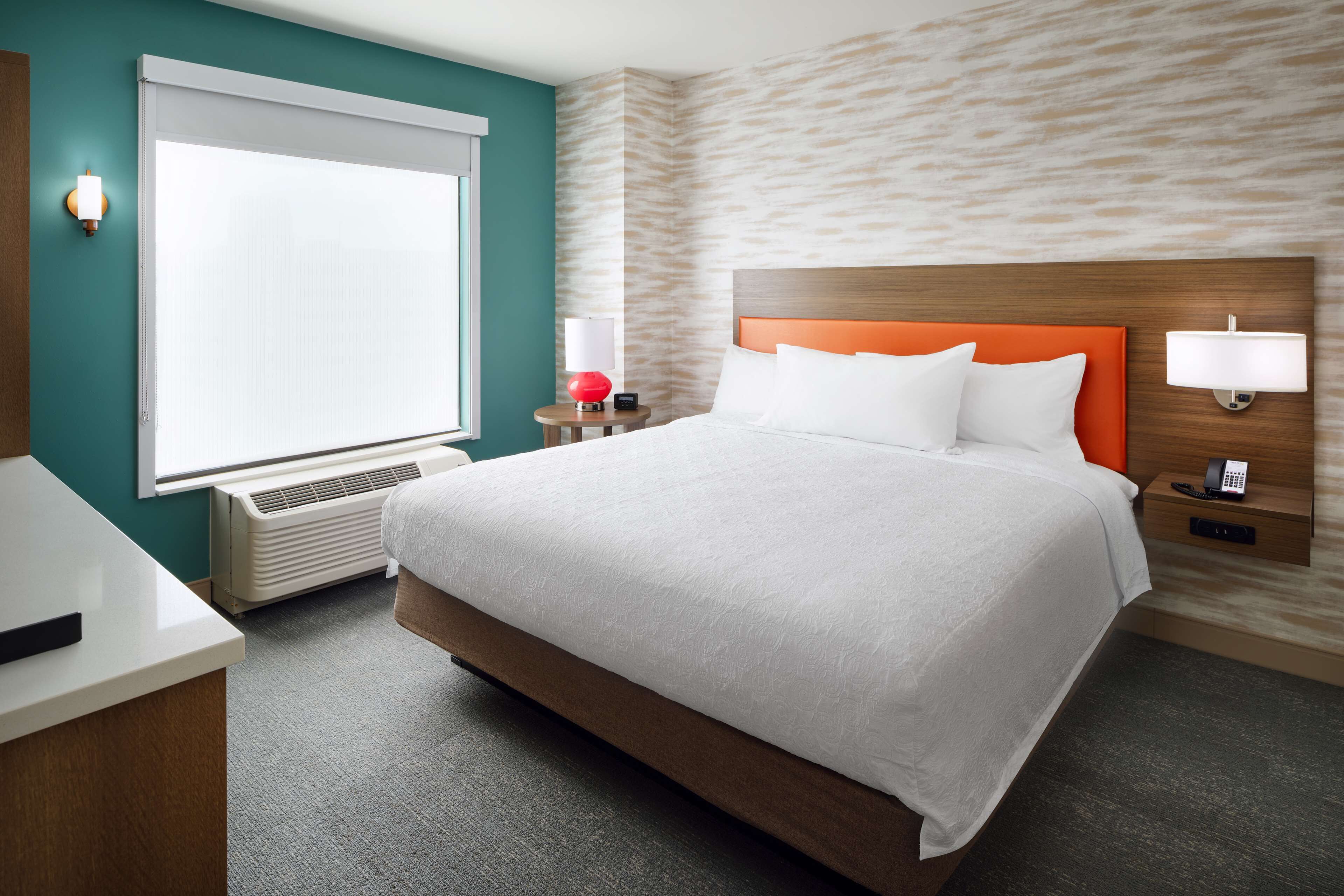 A hotel room with a large bed, a desk, a window with blinds, a wall-mounted lamp, and a phone on the nightstand.
