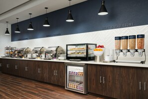 A self-service breakfast area with various food dispensers, a refrigerated display case, and a coffee station. - Home2 Suites by Hilton Towson (Towson)