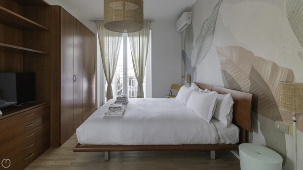 A bedroom with a wooden bed, a TV on a wooden cabinet, a bedside table with a lamp, and a window with curtains.