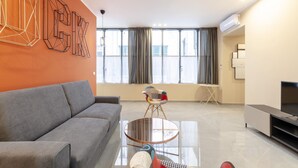 A modern living room with a grey sofa, a round glass coffee table, and a colorful chair. There is a flat-screen TV on a stand, a large window with curtains, and an air conditioning unit on the wall. - San Luca Apartments - Pallavicini by Wonderful Italy (Genoa)