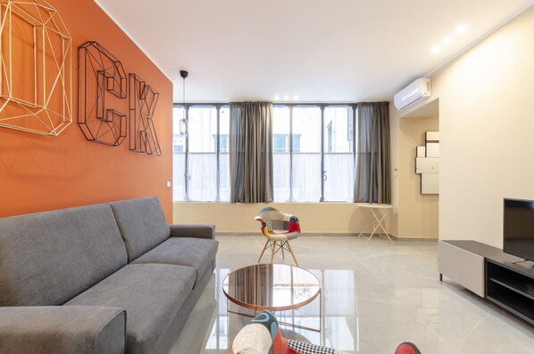 A modern living room with a grey sofa, a round glass coffee table, and a colorful chair. There is a flat-screen TV on a stand, a large window with curtains, and an air conditioning unit on the wall. - San Luca Apartments - Pallavicini by Wonderful Italy (Genoa)