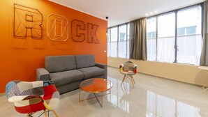 A modern living room with a grey sofa, a round glass coffee table, and a colorful armchair. The room features a large window with curtains and a wall with geometric wall art. - San Luca Apartments - Pallavicini by Wonderful Italy (Genoa)