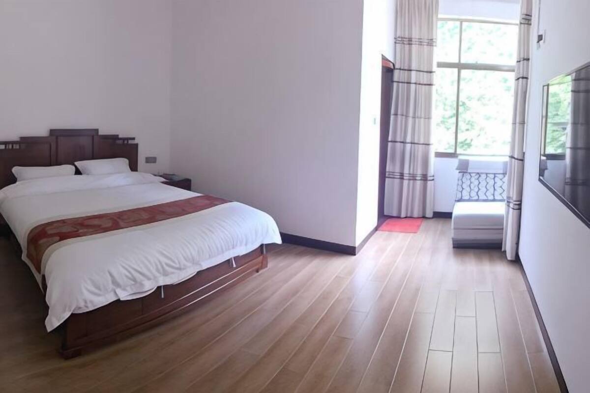 Una camera da letto con un letto di legno, un comodino, una finestra con tende e vista sul verde all'esterno.