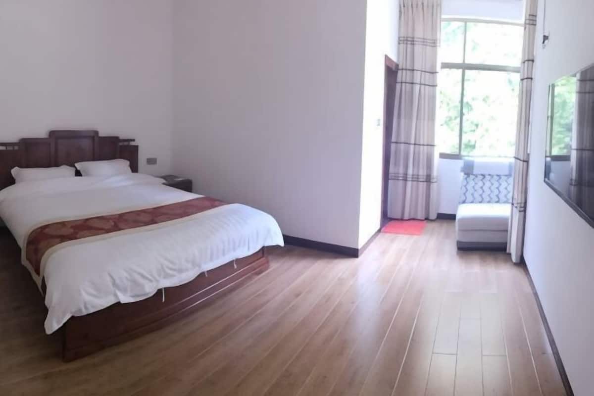 Camera da letto con un letto di legno, un comodino, una finestra con tende e vista sul verde.