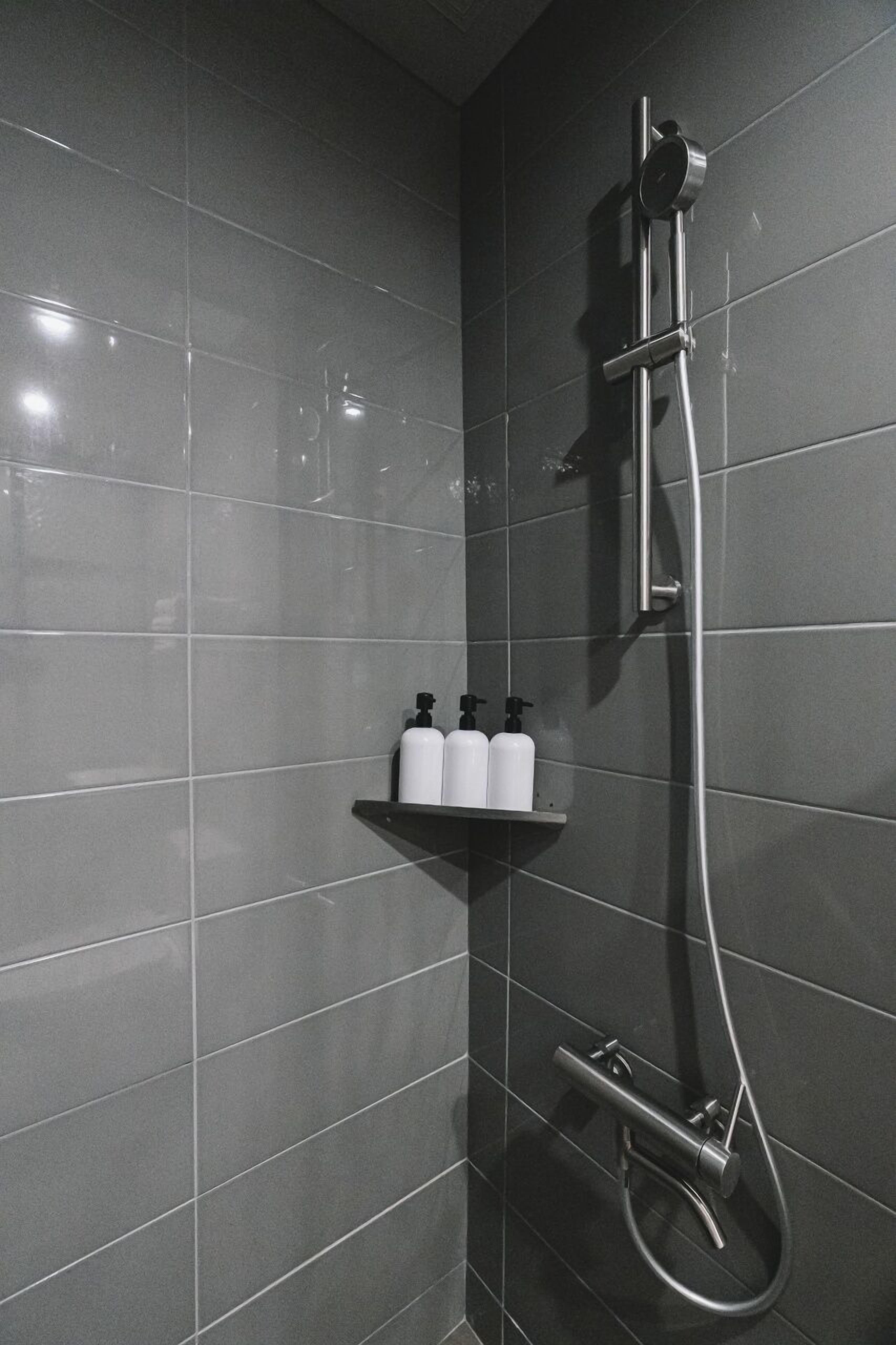 Ducha con cabezal de ducha de mano y grifo empotrado en la pared. Hay dos botellas blancas en un estante debajo de la ducha.