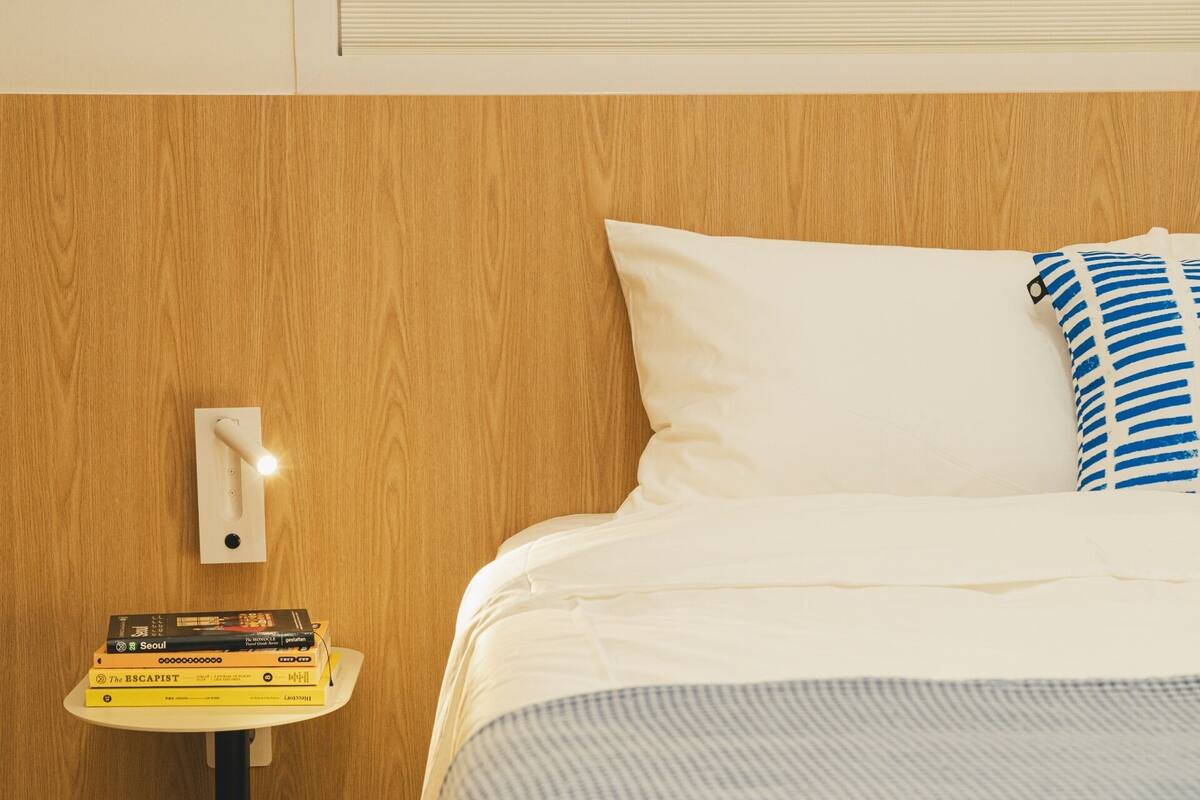 A neatly made bed with white and striped pillows, a bedside table with a stack of books, and a wall-mounted light switch.