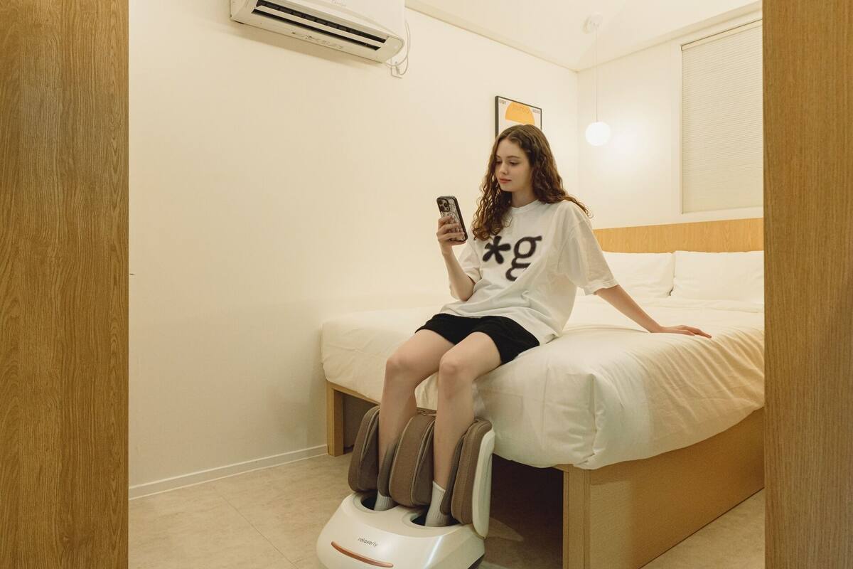 A person sitting on a bed in a room with a massage chair, a wall-mounted air conditioner, and a framed picture.