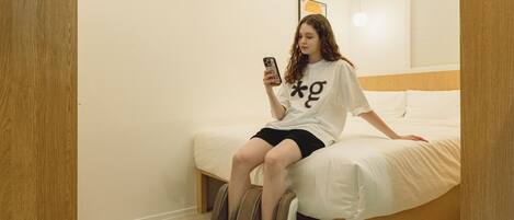 A person sitting on a bed in a room with a massage chair, a wall-mounted air conditioner, and a framed picture.