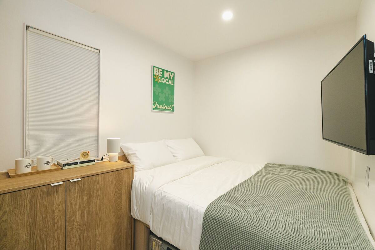 A bedroom with a bed, a bedside table with a lamp, a TV mounted on the wall, and a poster on the wall.