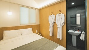 Habitación de hotel con cama, mesita de noche, perchero en la pared y baño con lavabo y espejo.