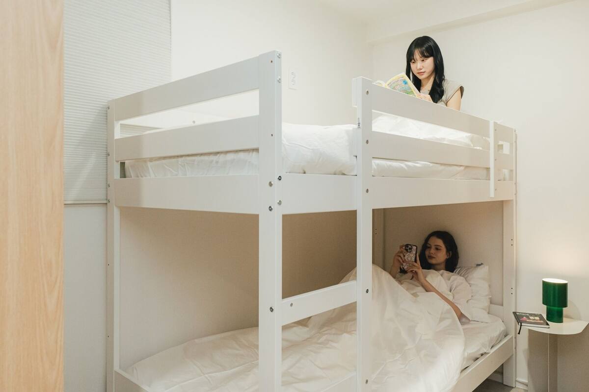 A bunk bed room with a window, a bedside table with a lamp, and a green object on the table.