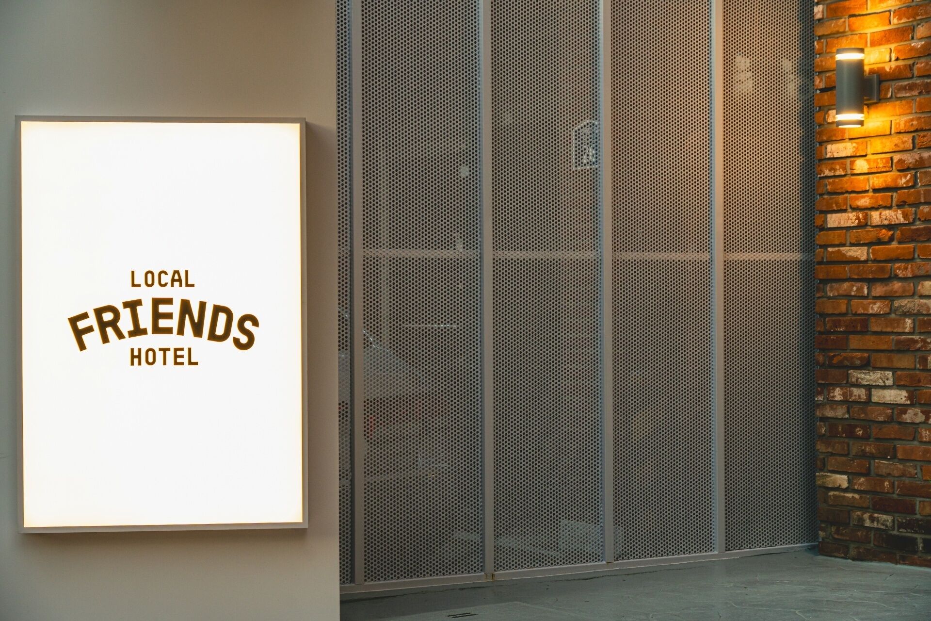 A sign for 'Local Friends Hotel' with a metal mesh wall and a brick wall with a light fixture.