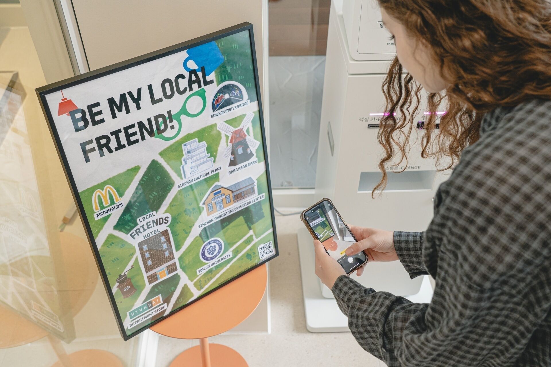 A person using a smartphone to interact with a digital map display titled "BE MY LOCAL FRIENDS".