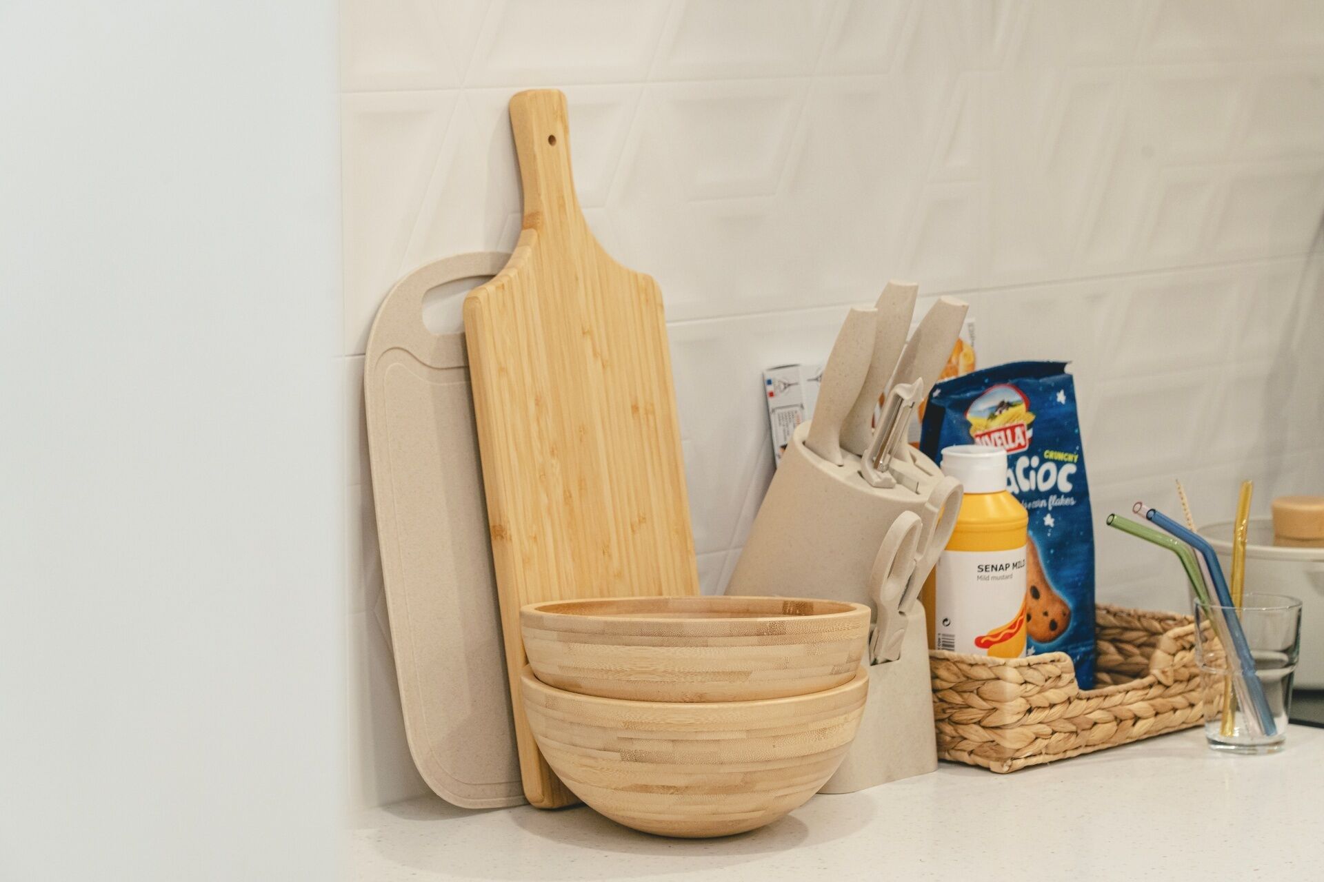 Una encimera de cocina con una tabla de cortar de madera, un cuenco de madera, una botella de Senap Milk, un paquete de Cruxy Flakes y una cesta tejida.