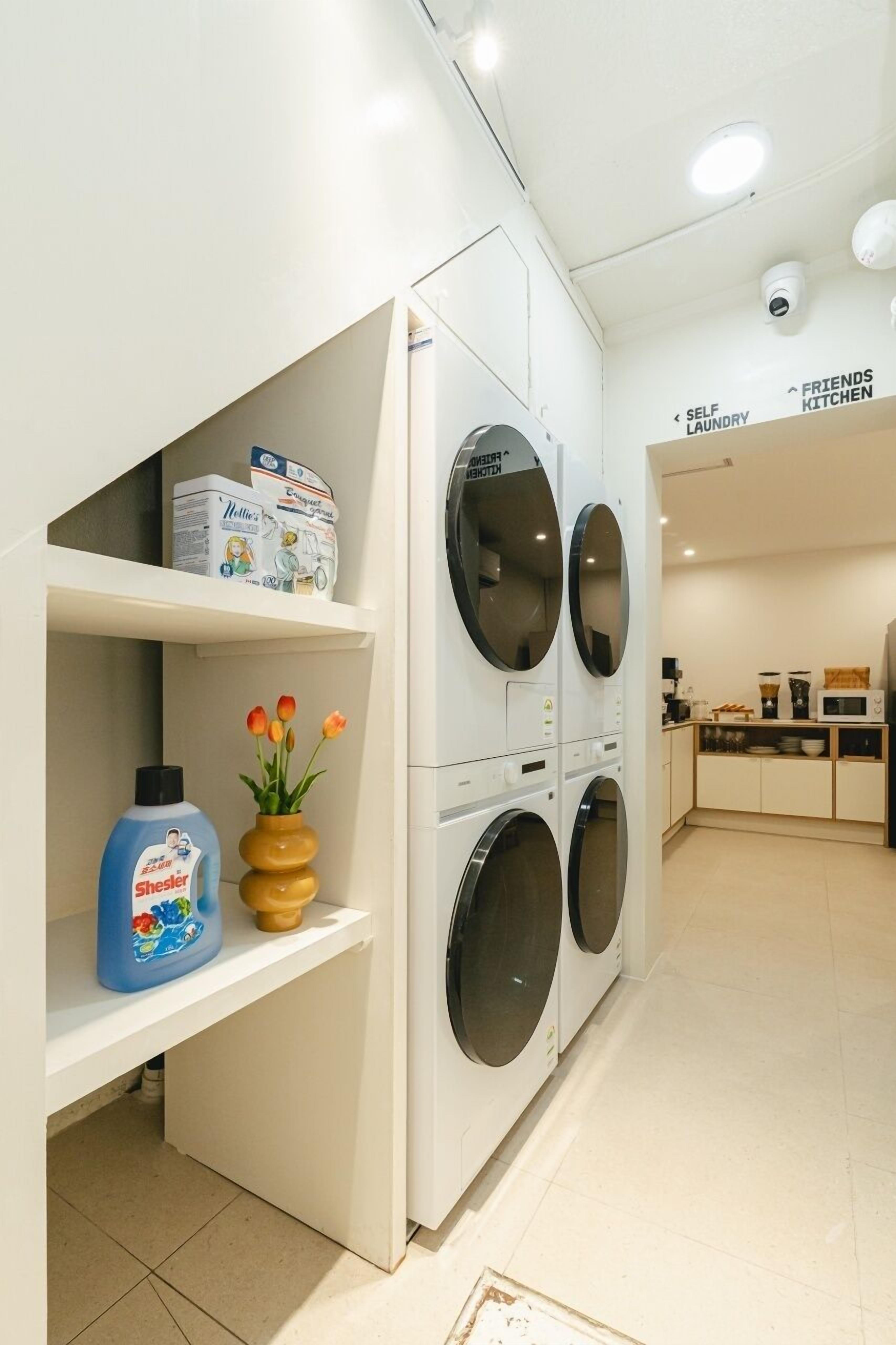 A laundry area with a washing machine, a shelf with detergent and a vase of flowers.