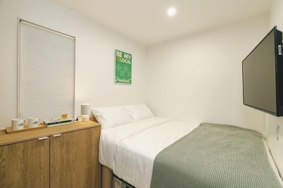 Une chambre à coucher avec un lit, une table de chevet avec une lampe, un téléviseur fixé au mur et une affiche accrochée au mur.