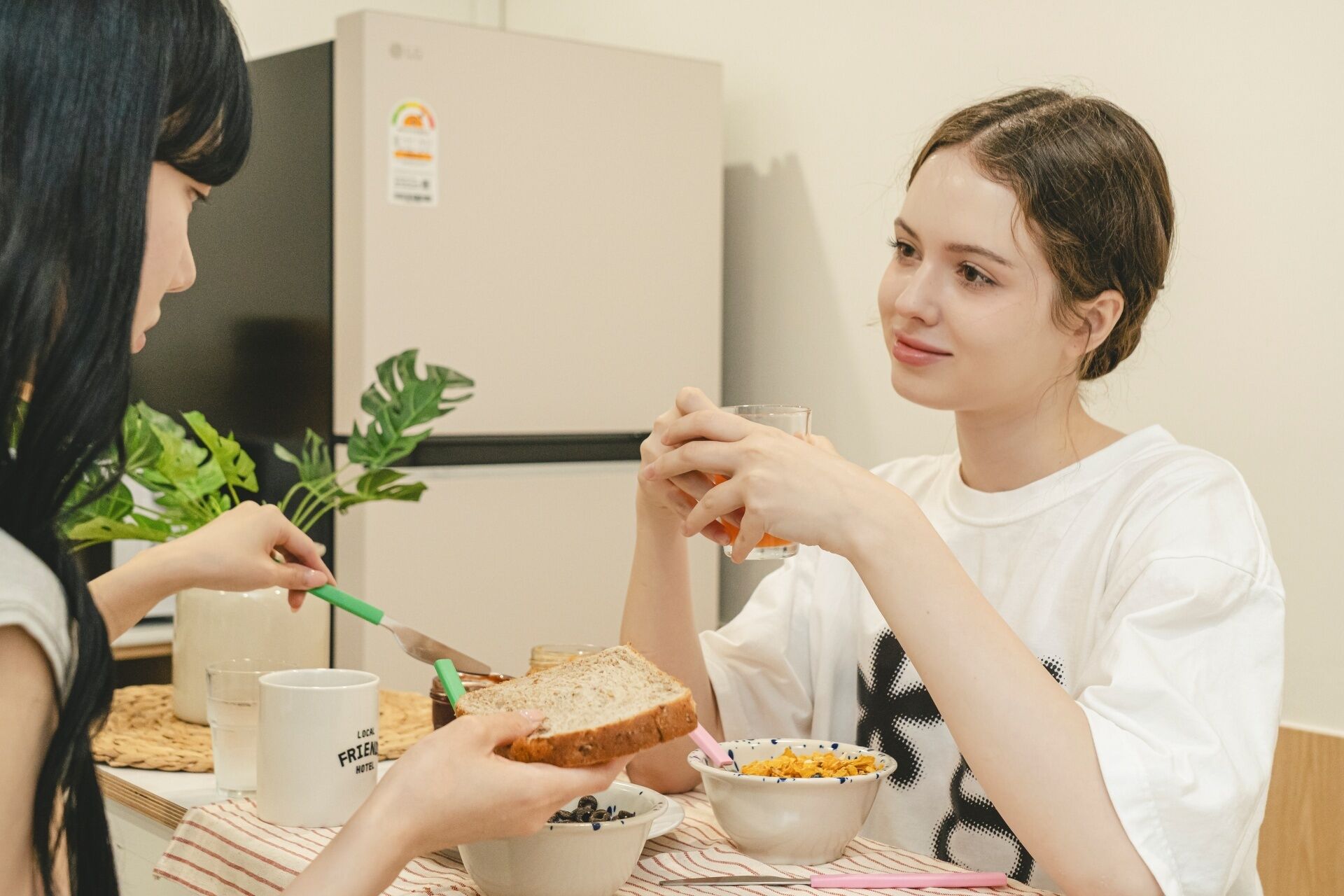 Two people sitting at a table with a bowl of cereal, a glass of juice, and a jar of jam.