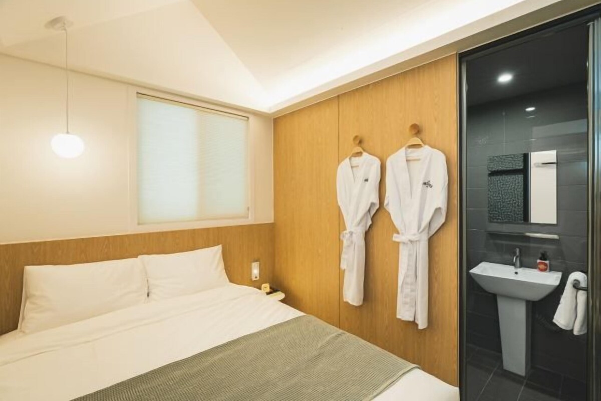A hotel room with a bed, a bedside table, a nightstand, a window with blinds, a wall-mounted robe rack, a glass shower enclosure, and a sink.