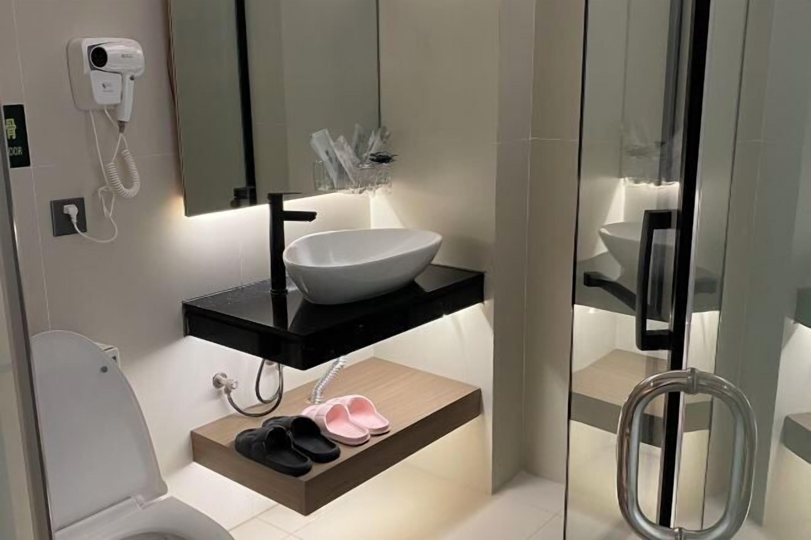 A modern bathroom with a white sink, black countertop, and a mirror above it. There is a hair dryer on the wall, a shelf with shoes, and a glass shower enclosure.