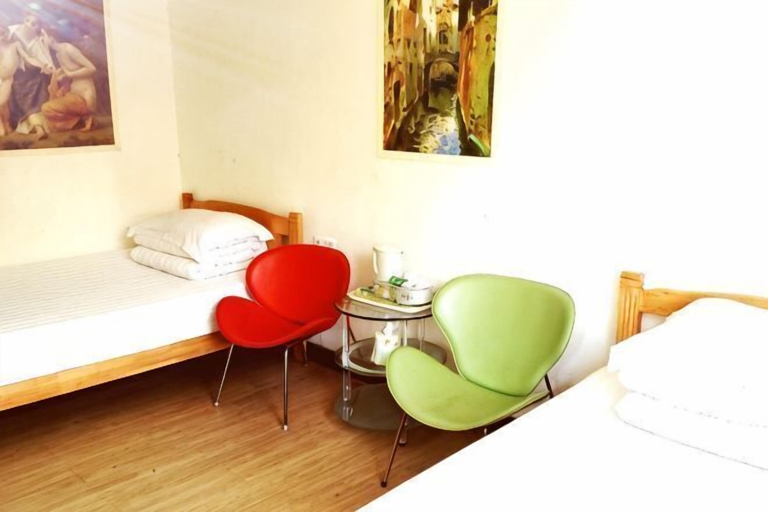 Ein Zimmer mit einem Bett, einem roten Sessel, einem grünen Sessel, einem kleinen Tisch mit einer Lampe und einem Bild an der Wand.