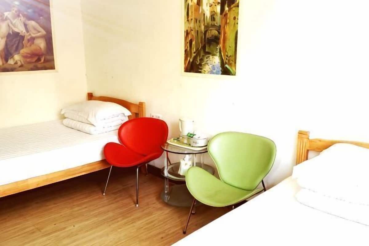 Una habitación con una cama, una silla roja, una silla verde, una mesita con una lámpara y un cuadro en la pared.