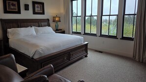 Un dormitorio ordenado con una cama grande, una mesita de noche, una lámpara y vistas a zonas verdes a través de las ventanas.
