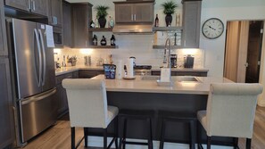 Uma cozinha moderna com geladeira de aço inoxidável, uma ilha central com banquetas e um relógio na parede.
