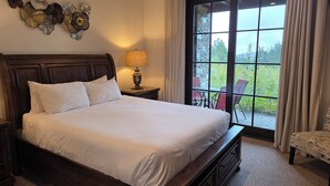 Una cama bien hecha con sábanas blancas, un cabecero de madera y una mesita de noche con una lámpara. Un ventanal con cortinas ofrece vistas al exterior.