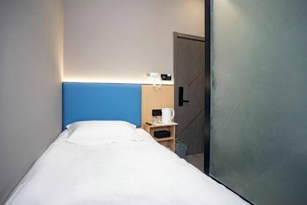 Quarto pequeno com uma cama de solteiro, cabeceira azul, mesa de cabeceira com chaleira branca e porta com puxador.
